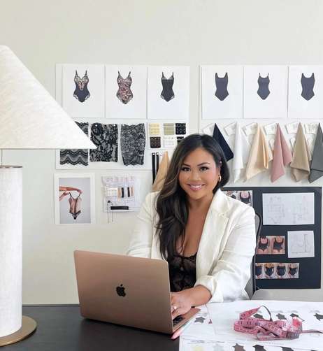 Designer working on a laptop, surrounded by lingerie sketches, fabric samples, and design tools.